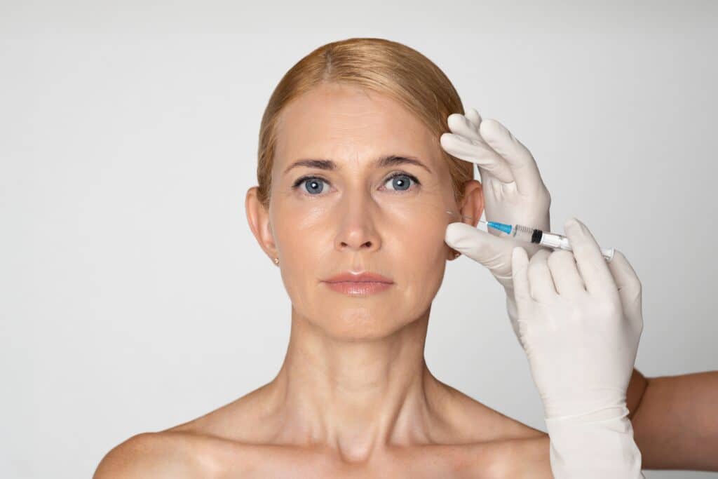 Attractive blond woman stares at camera, looking confident as a gloved hand injects Sculptra dermal filler in her cheek